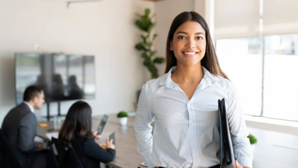 Woman holding binder with people working at desk behind her at office